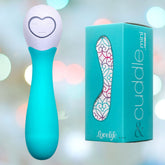 A turquoise electronic device, ideal for travel, showcases a heart-shaped control panel and is positioned next to its matching box labeled "cuddle mini" and "OhMiBod." The background is embellished with softly blurred lights in a spectrum of colors.